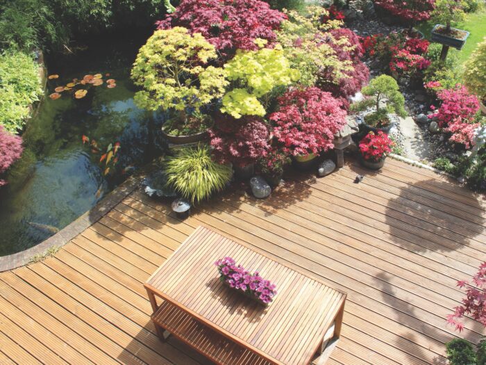 Photo showing a landscaped contemporary Japanese garden with a large expanse of pre-treated timber decking, providing a family space for outdoor furniture - a modern slatted table and benches.  The stained decking has been cut to form a curve around a large koi pond, while other features include Japanese maples, bonsai trees and oriental lanterns.